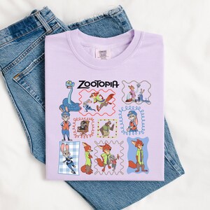 May include: A light purple t-shirt featuring the word "ZOOTOPIA" in black text. The shirt displays various cartoon character illustrations within decorative frames. The shirt is placed on top of a pair of blue denim jeans.