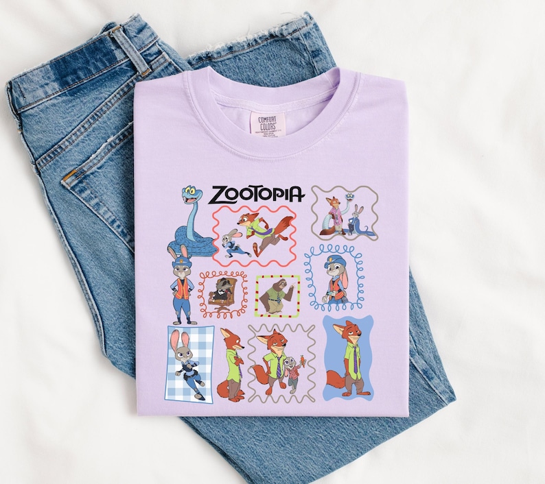 May include: A light purple t-shirt featuring the word "ZOOTOPIA" in black text. The shirt displays various cartoon character illustrations within decorative frames. The shirt is placed on top of a pair of blue denim jeans.