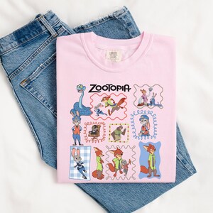 May include: A light pink t-shirt featuring the word "Zootopia" and various cartoon character illustrations. The shirt is folded over a pair of blue denim jeans. The shirt has a crew neck and a tag on the upper back.