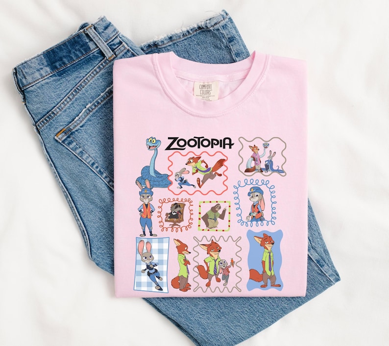 May include: A light pink t-shirt featuring the word "Zootopia" and various cartoon character illustrations. The shirt is folded over a pair of blue denim jeans. The shirt has a crew neck and a tag on the upper back.