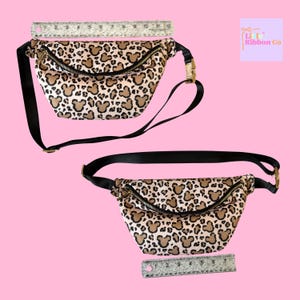 K&ouml;nnte beinhalten: Zwei Bauchtaschen mit einem beigen und braunen Mickey Mouse-Print. Jede Tasche hat einen verstellbaren schwarzen Riemen, eine goldfarbene Schnalle und einen schwarzen Rei&szlig;verschluss. Ein Lineal befindet sich in der N&auml;he jeder Tasche. Der Hintergrund ist rosa.