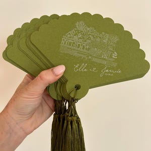 May include: A stack of olive green scalloped paper fans featuring a white line drawing of a building labelled The Great House, with the names Ella and Jamie and the date 29.08.26 printed below. Each fan has a matching olive green tassel attached.