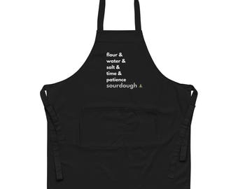 May include: Black apron with a large front pocket and neck strap. The apron features white text listing ingredients and steps for sourdough bread: flour, water, salt, time, patience, and sourdough.