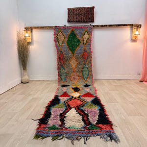 Puede incluir: Una alfombra rectangular larga y colorida con un patrón de diamantes en tonos grises, verdes, rojos y rosas. La alfombra tiene una textura peluda y bordes con flecos. Se exhibe sobre un suelo de madera de color claro.