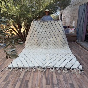 Peut inclure: Un grand tapis crème avec un motif géométrique en losange noir. Le tapis a un poil épais et texturé et une frange sur le bord inférieur. Le tapis est présenté à l'extérieur, une personne le tenant.