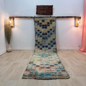 Puede incluir: Una alfombra larga y rectangular con un patrón geométrico en tonos azules, rosas y marrones. La alfombra se exhibe sobre un suelo de madera de color claro. Una pieza decorativa cuelga encima de la alfombra.