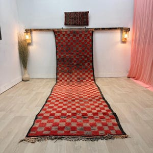 Puede incluir: Una alfombra larga y rectangular a cuadros rojos y beige con un borde negro. La alfombra se exhibe sobre un piso de madera clara. Una pieza rectangular más pequeña y a juego cuelga encima de la alfombra. La alfombra tiene flecos en la parte inferior.