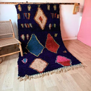 Peut inclure: Un tapis rectangulaire bleu marine profond à poils longs et épais, orné de grands motifs en forme de losanges aux couleurs variées, notamment crème, bleu sarcelle, rouge et rose. Le bas du tapis est agrémenté de franges nouées.