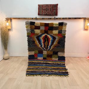 Puede incluir: Una colorida alfombra rectangular con un diseño geométrico. La alfombra presenta una forma de diamante central en azul, rojo y amarillo, rodeada por un mosaico de cuadrados de varios colores. La alfombra tiene un borde con flecos.