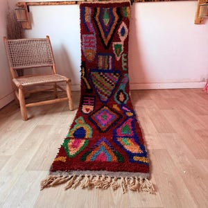 Peut inclure: Un long tapis rectangulaire de couleur rouge foncé orné de motifs géométriques multicolores en bleu, vert, jaune et rose. Le tapis présente un poil épais et texturé ainsi que des franges de couleur beige clair à l'une de ses extrémités.