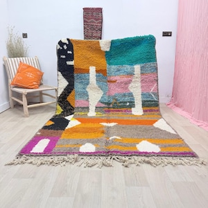 Peut inclure: Un tapis marocain coloré avec un motif patchwork géométrique. Le tapis présente une variété de couleurs, notamment orange, jaune, rose, bleu et blanc. Il a une bordure à franges et est présenté sur un sol en bois clair.