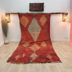 Puede incluir: Una alfombra roja grande con un patrón de diamantes en naranja y blanco. La alfombra está colgada de una viga de madera con dos pequeñas lámparas a cada lado. Una almohada más pequeña con estampado está encima de la alfombra.