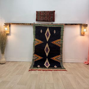 Puede incluir: Una alfombra negra y multicolor con un diseño geométrico y de diamantes. La alfombra está colgada de una viga de madera con dos apliques. Una almohada más pequeña con estampado está encima de la alfombra.