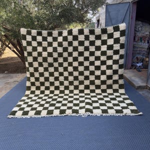 Peut inclure: Un grand tapis rectangulaire à motif de damier vert olive et crème. Le tapis est fait d'une matière épaisse et texturée et a un bord frangé. Le tapis est exposé à l'extérieur.
