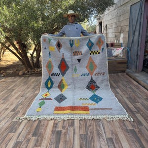 May include: A gray rug with a geometric diamond pattern in various colors, including red, orange, yellow, and blue. The rug has a fringe along the bottom edge and is displayed outdoors.