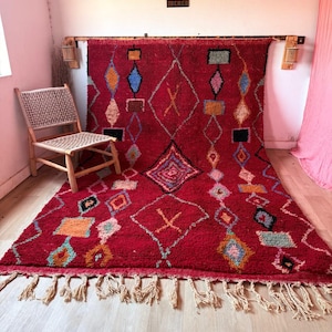 Peut inclure: Un grand tapis rectangulaire rouge profond orné d'un motif géométrique composé de losanges et de carrés bleus, orange, jaunes et noirs. Le tapis présente un poil épais et texturé, terminé par de longues franges nouées de couleur crème.
