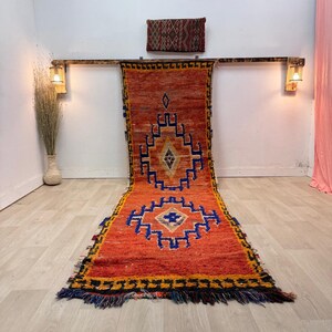 Puede incluir: Una alfombra larga y rectangular con un vibrante fondo naranja y patrones geométricos en azul, amarillo y negro. La alfombra tiene un borde con flecos y se exhibe sobre un suelo de madera clara. La alfombra es un elemento decorativo para el hogar.