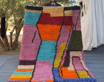 May include: A large, colorful rug with a geometric design. The rug features a variety of colors, including pink, orange, blue, green, and yellow. It has a shaggy texture and a fringe along the bottom edge. The rug is being held up by a person.