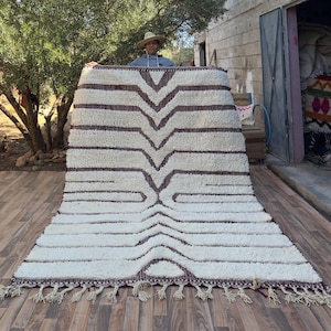 May include: A large, rectangular off-white rug with a geometric pattern in dark brown. The rug has a thick, textured pile and a fringe along the bottom edge. The design features a series of chevrons and horizontal lines.