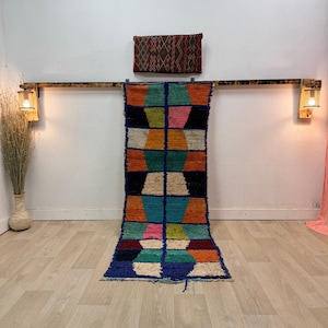 Puede incluir: Una colorida alfombra de corredor geométrica con un borde azul. La alfombra presenta un patrón de formas naranjas, turquesas, rosas y negras. Un cojín decorativo con un diseño estampado cuelga sobre la alfombra. La alfombra está sobre un suelo de madera clara.