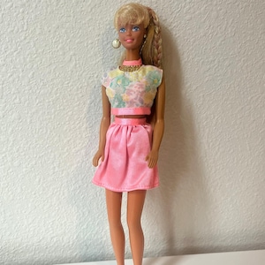 Puede incluir: Muñeca vintage con cabello rubio peinado en una trenza lateral. Viste un top corto con estampado floral, una falda rosa y pendientes dorados. El atuendo de la muñeca se completa con una gargantilla rosa y un cinturón a juego.
