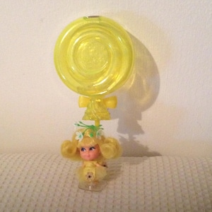 May include: A vintage yellow plastic doll with a large lollipop-shaped top. The doll has blonde hair, a floral crown, and a yellow dress. The lollipop is attached to the doll with a bow. The doll is standing on a clear base.