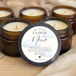 May include: Five small, brown glass jar candles with white wax and wicks. One candle has a lid with the text "WE'RE ON CLOUD Nine" and a cloud graphic. The background includes a blanket and baby booties.