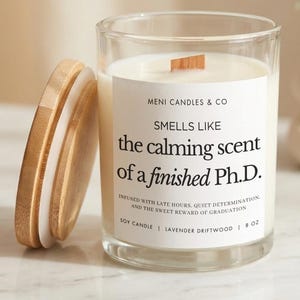 May include: A clear glass candle jar with a wooden lid, containing a white candle. The label reads "Smells Like the calming scent of a finished Ph.D." The candle is 8 oz and has a lavender driftwood scent.