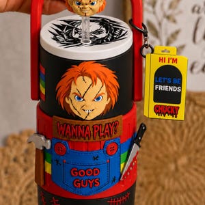 May include: A black tumbler featuring a Chucky character topper, a straw, and a red handle. The body displays a Chucky face, the text WANNA PLAY?, a blue Good Guys overall pocket, a hammer, a knife, and red stitches. A yellow tag reads HI I'M LET'S BE FRIENDS CHUCKY.