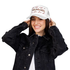 May include: White trucker hat with the word "MOM MOM MOM MOM" in a floral design across the front. The hat has a mesh back and a curved brim. The hat is worn by a person wearing a black denim jacket.