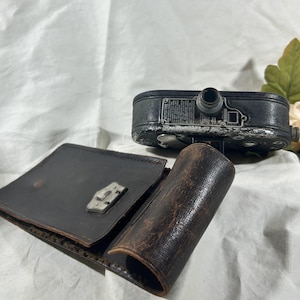 May include: An antique black and silver Keystone movie camera with a leather case and lens cover. The camera has a silver lens and several dials and buttons. The leather case is open, revealing a silver clasp.