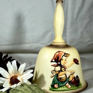May include: Cream-colored ceramic bell with a long handle. The bell features a painted illustration of a girl in a green dress and brown headscarf, holding a basket. The bell has a brown rim and is decorated with flowers.