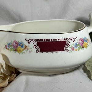 May include: White ceramic gravy boat with a gold rim and a curved handle. The boat features a burgundy rectangular design with floral accents in pink, blue, and yellow. A classic piece for serving sauces and gravies.