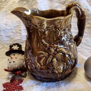 May include: A brown ceramic pitcher with a detailed relief design featuring grapes and animals. The pitcher has a curved handle and a spout for pouring. A snowman ornament and a red snowflake are next to the pitcher.