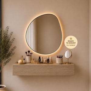 Peut inclure: Un miroir mural de forme organique irrégulière avec rétroéclairage LED intégré, fixé au-dessus d'une console flottante en bois clair. L'étagère contient des flacons cosmétiques, des pinceaux et un petit miroir de courtoisie. Texte : READY TO MOUNT FULLY ASSEMBLED.