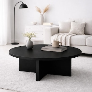 Könnte beinhalten: Ein runder, schwarzer Couchtisch mit massivem Sockel. Der Tisch befindet sich in einem Wohnzimmer mit einem weißen Sofa, einem Teppich und einer schwarzen Stehlampe. Eine Vase mit Blumen und eine Tasse stehen auf dem Tisch.