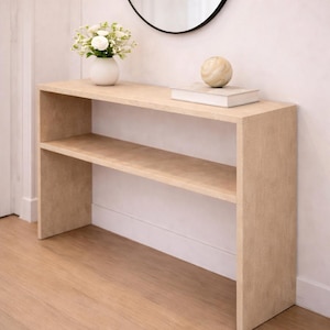 May include: A light-colored wooden console table with two shelves. A small white vase with white flowers, a book, and a decorative sphere are on the top shelf. A round black mirror hangs on the wall above the table.