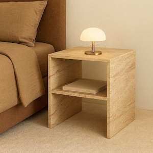 May include: A neutral-toned bedroom scene featuring a bedside table with a small lamp and a book. The table is made of a light-colored, textured material. The lamp has a white, dome-shaped shade and a brass base. A bed with a brown headboard and bedding is in the background.