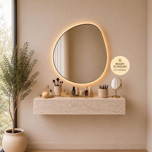 May include: A modern vanity setup featuring a large, irregularly shaped mirror with a warm, glowing border. The mirror hangs above a stone-textured floating shelf holding makeup and accessories. A potted plant and a small round mirror complete the elegant design.