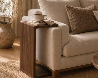 Könnte beinhalten: Ein C-förmiger Beistelltisch aus dunklem Holz neben einem beigen Polstersofa. Der Tisch hat eine flache Oberfläche mit einer Keramiktasse und einem Buch sowie einen stabilen rechteckigen Fuß, der unter den Rahmen des Sofas geschoben wird.