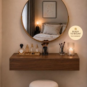 Op de afbeelding: Een zwevende kaptafelplank van donker hout met daarboven een ronde spiegel met gouden rand. Op de plank staan parfumflesjes, make-upkwasten en een kaars. Daaronder staat een crèmekleurige, drielaagse cilindrische kruk met een gouden voet. Tekst: READY TO MOUNT.