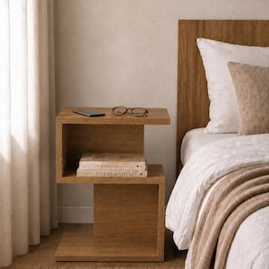 May include: A wooden nightstand with a minimalist design, featuring two shelves. The top shelf holds a pair of eyeglasses and a smartphone. Two books are stacked on the lower shelf. A bed with white linens and a brown throw blanket is next to the nightstand.