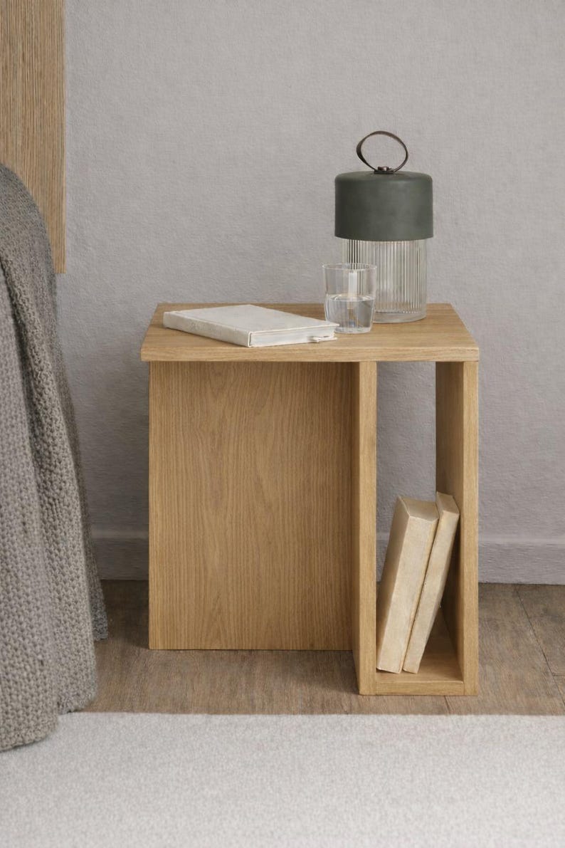 May include: A light oak bedside table with a rectangular top and an open shelf. A green and clear glass lamp, a glass of water, and a book are on the table. Two books are in the open shelf.