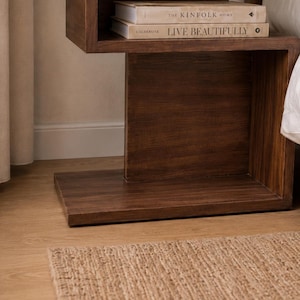 May include: A dark brown wooden nightstand with a built-in shelf holding two books. The books are titled "THE KINFOLK HOME" and "LIVE BEAUTIFULLY". A woven rug is on the floor in front of the nightstand.