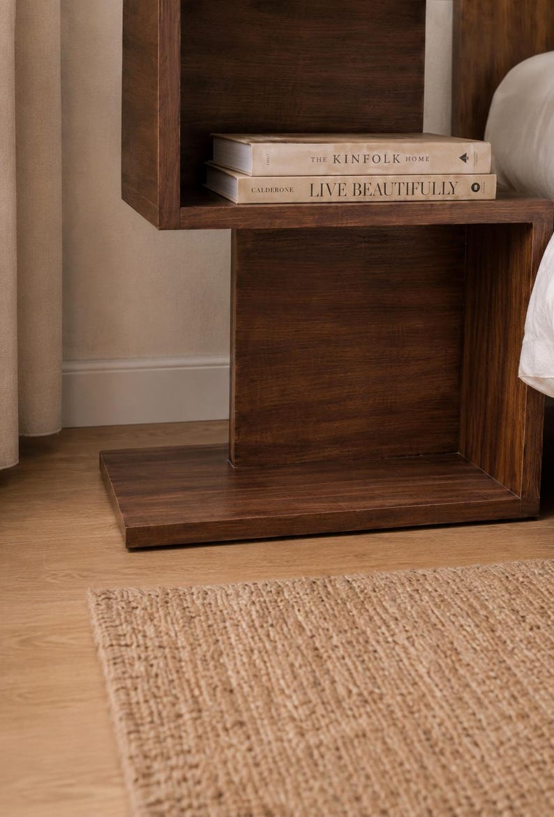 May include: A dark brown wooden nightstand with a built-in shelf holding two books. The books are titled "THE KINFOLK HOME" and "LIVE BEAUTIFULLY". A woven rug is on the floor in front of the nightstand.