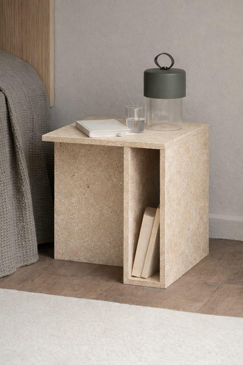 May include: A light beige, modern nightstand with a built-in shelf. A glass of water, a book, and a decorative lamp with a dark green top and a clear, ribbed glass base sit on top. Two books are in the shelf.