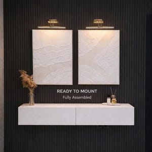 May include: Two textured, off-white wall art pieces with gold-toned lighting fixtures above. A white floating cabinet with two drawers is below the art. The text "READY TO MOUNT Fully Assembled" is visible. A vase of dried flowers and decorative items are on the cabinet.