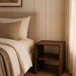 May include: A dark brown wooden nightstand with two shelves stands next to a bed with beige bedding and pillows. A brown throw blanket and a woven rug are at the foot of the bed. A framed landscape painting hangs on the wall.
