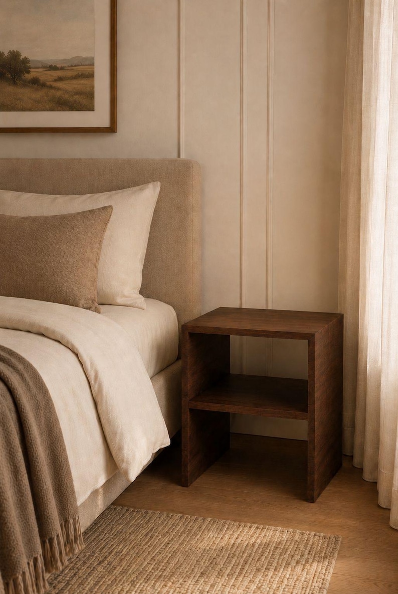 May include: A dark brown wooden nightstand with two shelves stands next to a bed with beige bedding and pillows. A brown throw blanket and a woven rug are at the foot of the bed. A framed landscape painting hangs on the wall.