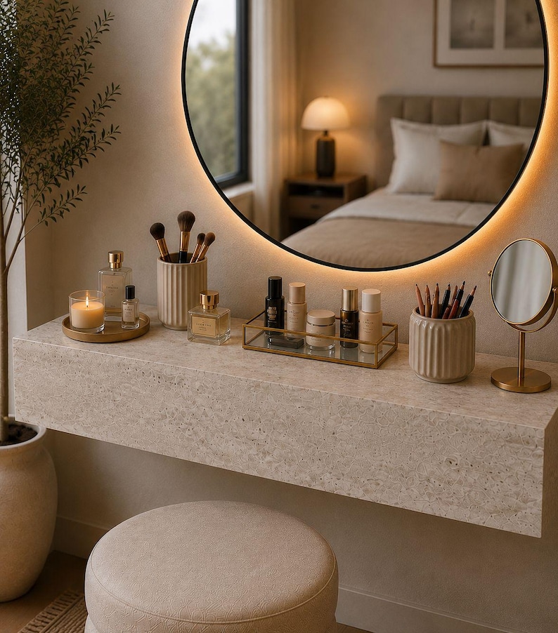 May include: A neutral-toned vanity with a large round mirror and a variety of beauty products. The vanity features a stone-like surface with a stool in front. Makeup brushes, perfumes, and skincare items are arranged on the vanity, creating a serene and organized space.